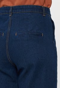 Mørkeblå denimbukser med en glat tekstur, der har en baglomme med diskret syning og en figursyet taljedesign.