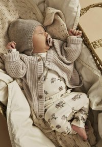 Cardigan beige tricoté avec des boutons en bois, bodysuit crème à motif floral et pantalons, bébé dans un panier tissé douillet avec des couvertures douces.