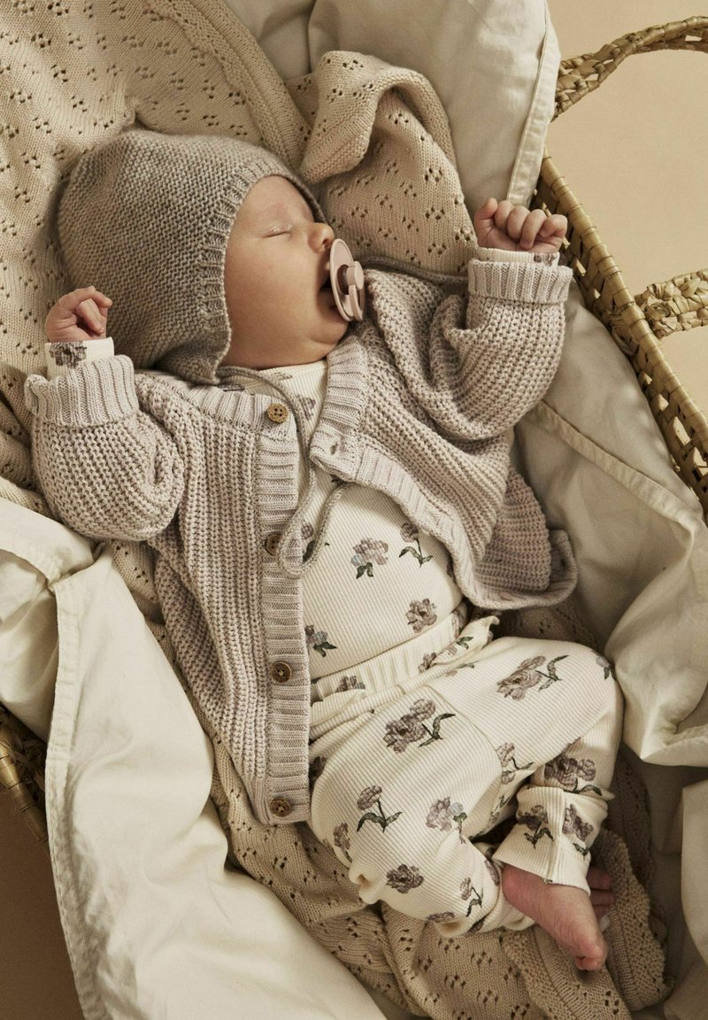 Cardigan beige tricoté avec des boutons en bois, bodysuit crème à motif floral et pantalons, bébé dans un panier tissé douillet avec des couvertures douces.