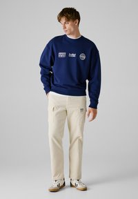 Sweatshirt bleu marine avec logos imprimés, associé à un pantalon cargo crème avec poches et une étiquette. Des baskets avec des accents noirs complètent le look.