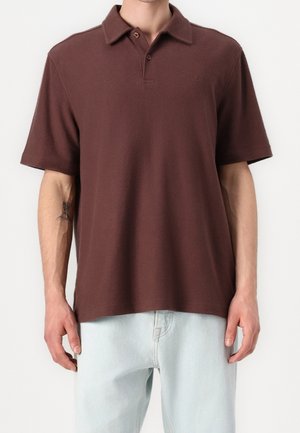 Brown polo shirt with a soft texture, short sleeves, and a three-button placket. Worn with light blue denim trousers.
