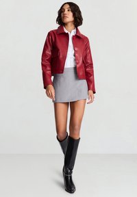 Veste courte en similicuir rouge avec deux poches frontales, associée à une mini-jupe grise et des bottes noires montant au genou, mettant en valeur des textures lisses.