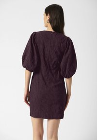 Mujer vestida con un vestido texturizado de un púrpura oscuro con mangas abullonadas hasta el codo, de pie de espaldas contra un fondo claro y sencillo.