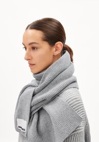 Écharpe grise à côtes enroulée autour du cou, avec une étiquette sur un côté. Le tissu semble doux et texturé, complétant un pull rayé.