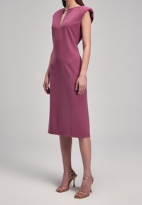 Robe cintrée à manches courtes en forme de casquette, de couleur mauve foncé, avec un décolleté en V et des coutures verticales. Associée à des talons hauts dorés à brides. Texture lisse.