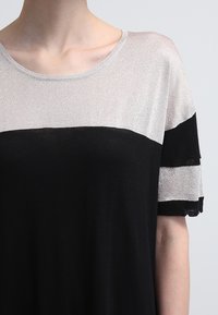 Femme portant un haut en tricot à manches courtes avec une partie supérieure en argent métallique et une partie inférieure noire, ainsi que des rayures sur les manches, sur un fond clair.