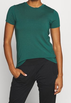 Person wearing a fitted short-sleeve green t-shirt and loose black pants with one hand in the pocket against a plain background.