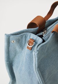 Lysblå denim tote bag med brune stofhåndtag. Har sølvhardware, lynlåslukning og tekstureret stofmønster.