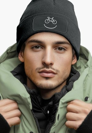 Black knit beanie with a bicycle and smile graphic on a rectangular patch, paired with a green puffy jacket over a black interior layer.