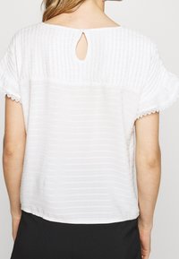 Bílá blůza s texturovaným pruhovaným vzorem, krátkými řasenými rukávy a výstřihem na zádech s knoflíkovým zapínáním. Rukávy zdobí krajkový lem.