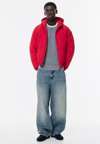 Jachetă roșie căptușită cu glugă, pulover gri cu nervuri și blugi lejere din denim albastru. Pantofi negri completează ținuta casual.