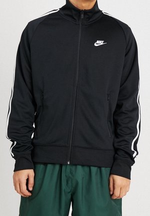 Personne portant une veste zippée Nike noire avec des bandes blanches sur les manches et un short vert foncé, debout sur un fond clair.