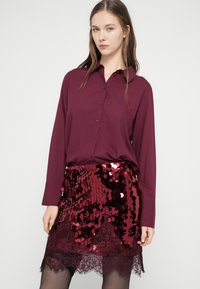 Bordowa koszula na guziki z długimi rękawami, zestawiona z spódnicą ozdobioną cekinami i koronkowym wykończeniem na dole.