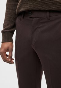 Calças castanho-escuras feitas à medida com textura suave, apresentando bolsos frontais e fecho com botão, combinadas com um suéter castanho texturizado.