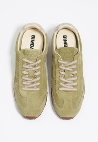 Oliwkowe zamszowe sneakersy z jasnobrązowymi gumowymi podeszwami, z teksturowanymi sznurowadłami i przeszywaną konstrukcją. Logo wytłoczone na języku.