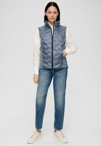 Gepolsterte graue Weste mit hohem Kragen und Reißverschluss. Verfügt über seitliche Taschen, strukturiere Vorderseite und glatte Seiten. Getragen über einem weißen Hemd und blauen Jeans.