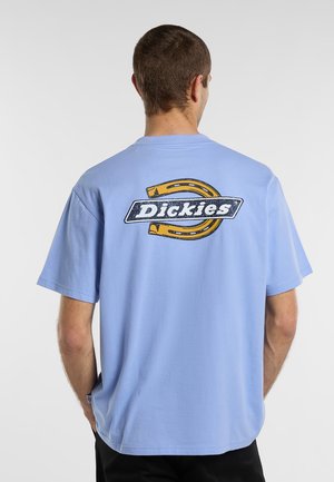 Man som står med ryggen mot, iklädd en ljusblå t-shirt med en stor gul hästsko och "Dickies"-logotyp på ryggen.