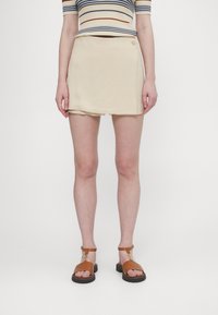 Beige skort med en enkel design som har en knapprad. Bärs tillsammans med bruna sandaler med remmar, mot en vit bakgrund.