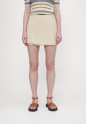 Beige skort met een eenvoudig ontwerp, voorzien van een knoopdetail. Gecombineerd met bruine sandaaltjes met banden, op een witte achtergrond.