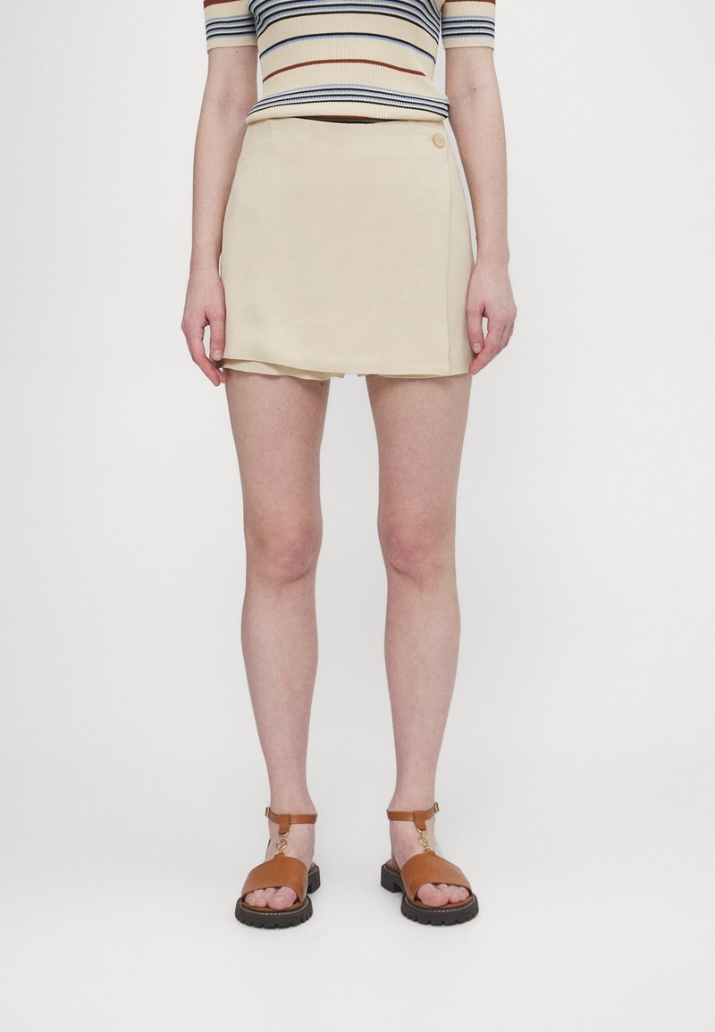 Beige skort med en enkel design som har en knapprad. Bärs tillsammans med bruna sandaler med remmar, mot en vit bakgrund.