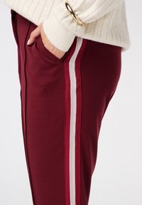 Pantalons de survêtement bordeaux avec des rayures blanches sur les côtés, présentant une texture douce et des poches, conçus avec une coupe fuselée pour le confort.