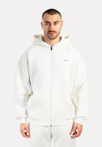 Muž s krátkými vlasy a plnovousem, oblečený v bílé mikině na zip s předními kapsami a odpovídajících bílých kalhotách, proti jednobarevnému pozadí.