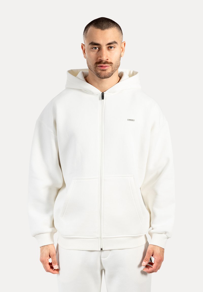 Muž s krátkými vlasy a plnovousem, oblečený v bílé mikině na zip s předními kapsami a odpovídajících bílých kalhotách, proti jednobarevnému pozadí.