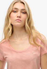 Jasnoróżowa koszulka z krótkim rękawem o okrągłym dekolcie, wykonana z miękkiego, teksturowanego materiału. Posiada subtelnie wyhaftowane logo na piersi.