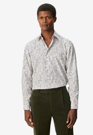 Uomo che indossa una camicia bianca a maniche lunghe con un piccolo motivo floreale colorato e pantaloni verde scuro su sfondo neutro.
