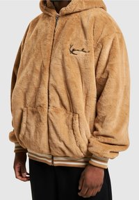 Kurtka polarowa z kapturem w jasnobrązowym kolorze o miękkiej strukturze, z prążkowanymi mankietami i krawędzią oraz przednimi kieszeniami. Posiada mały wyszywany wzór.