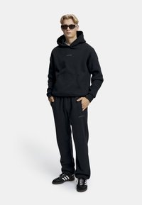 Mustapaita etutaskulla ja logolla, yhteensopivat mustat hikoiluhousut, mustat lenkkitossut, joissa on valkoiset raidat, malli käyttää aurinkolaseja.