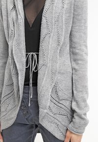 Grå stickad cardigan med öppen front, som har strukturerade vågmönster och fickor. Kombinerad med en svart topp och mörkgrå byxor.