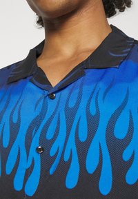 Koszula z krótkim rękawem z stopniowym niebieskim gradientem i czarnymi wzorami płomieni. Czarny kołnierz z ozdobnym guzikiem przy dekolcie.