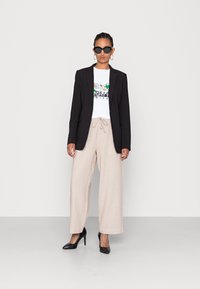 Svart kavaj, vit grafisk t-shirt, beige vida byxor, svarta högklackade skor, stora svarta solglasögon och runda örhängen.