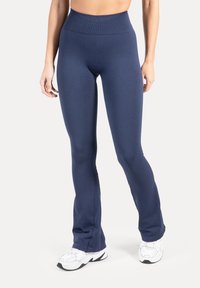 Frau trägt hoch taillierte, marineblaue figurbetonte Leggings und weiße Sportschuhe, steht vor einem schlichten hellen Hintergrund.