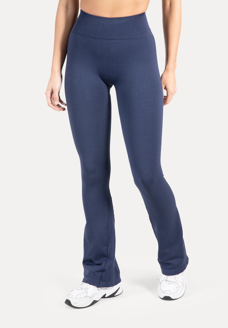 Frau trägt hoch taillierte, marineblaue figurbetonte Leggings und weiße Sportschuhe, steht vor einem schlichten hellen Hintergrund.