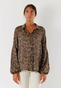 Blusa floreale marrone con maniche lunghe e puffate, chiusura a bottoni e vestibilità rilassata. Presenta un mix di motivi floreali verdi, viola e bianchi.