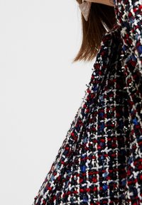 Veste en tissu tweed tissé présentant un motif multicolore avec des fils rouges, blancs et bleu marine, ayant une surface texturée et un léger éclat.