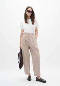 Beige pleated trousers with a high waist, paired with a plain white t-shirt and black loafers; holds a dark brown bag and wears black sunglasses.