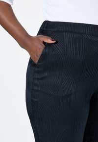 Schwarze, strukturierte Hose mit einem dezenten Wellenmuster, elastischem Bund und Seitentasche. Der Stoff hat einen sanften Glanz.