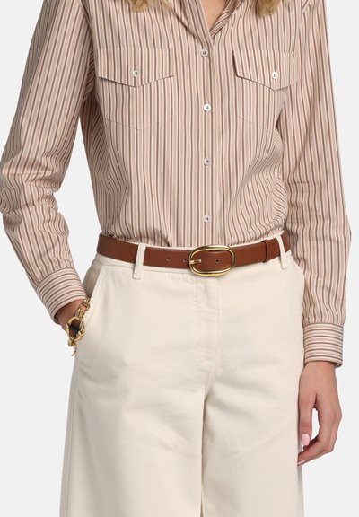 Žena nosi svetlobéžovú a bielo pruhovanú košeľu zapnutú na gombíky, zastrčenú do krémových nohavíc s hnedým opaskom a zlatým náramkom na ľavom zápästí.
