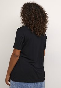 Black cotton t-shirt with short sleeves, relaxed fit, and a rounded hem. Worn with light denim skirt. Curly hair visible at the back.