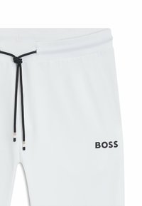 Witte joggers met een trekkoord in de taille, voorzien van zwarte accenten en het woord "BOSS" gedrukt op het rechteronderbeen. Glad van textuur.