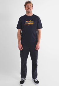 Homme debout avec les bras détendus, portant un t-shirt graphique noir, un pantalon sombre et des baskets noir et blanc, devant un fond blanc uni.