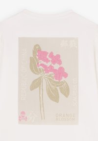 Weißes T-Shirt mit einem gedämpften, beigefarbenen floralen Grafikdesign, das rosa Blumen, Text in verschiedenen Schriftarten und einen rosa Akzent in Form eines Schädels zeigt.