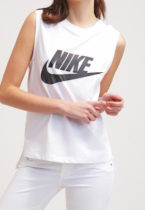 Femme portant un haut Nike blanc sans manches et un pantalon blanc, les mains jointes devant, sur fond uni.
