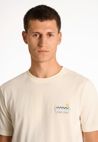 Vaaleanbeige lyhythihainen t-paita, jossa on brodeerattu aaltojen ja auringon kuva, ja tekstinä "SUN OF A BEACH" päällä.