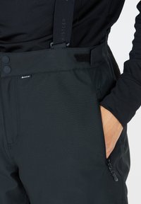 Zwarte waterdichte skibroek met verstelbare banden, twee knopen in de taille en een zijrits zak. Glad textuur met subtiele diagonale lijnen.