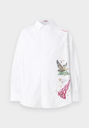 Homme portant un pantalon noir ample, des baskets noir et blanc, et une chemise blanche boutonnée avec des motifs d'aigle et de drapeau sur le côté.