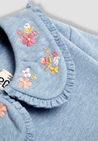 Denim modrá látka s volánovým golierom a vyšívanými kvetmi v ružovej, bielej a zlatej farbe; obsahuje zlaté hviezdy.